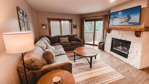 A living room with a sectional sofa, a coffee table, a fireplace, and a large TV mounted on the wall.