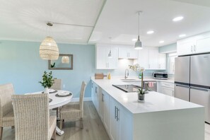 Une cuisine moderne avec des meubles blancs, un grand îlot central et un coin repas doté de chaises en osier.