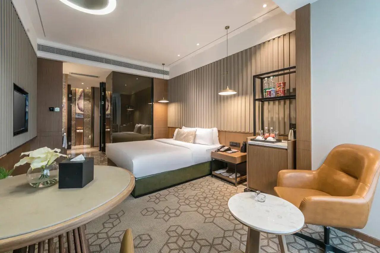 Habitación de hotel moderna con cama, mesita de noche, lámpara, silla y una pequeña mesa con un florero.