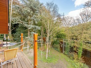 A wooden deck with wicker chairs, a glass railing, and a view of a river and trees.