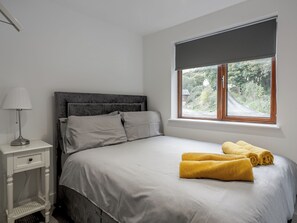 Ein Schlafzimmer mit Bett, Nachttisch, Lampe und einem Fenster mit Blick ins Grüne.