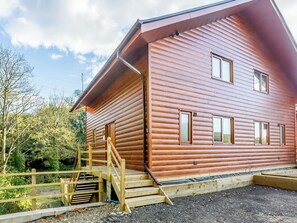 Eine Blockhütte mit einer Holztreppe, die zu einer Tür führt, umgeben von Bäumen und Kies.