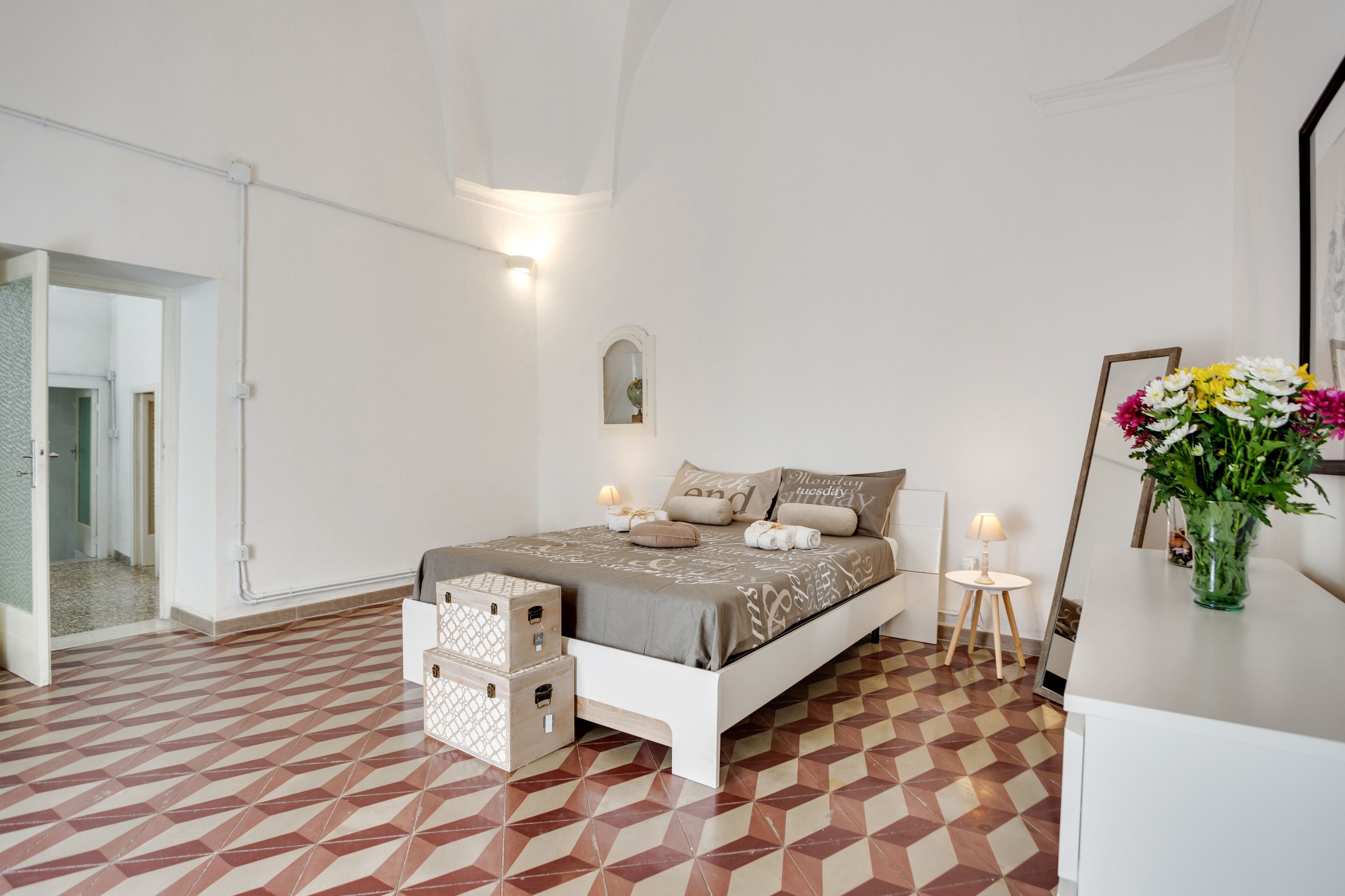 A bedroom with a bed, bedside lamps, a mirror, a vase with flowers, and a geometric patterned floor.