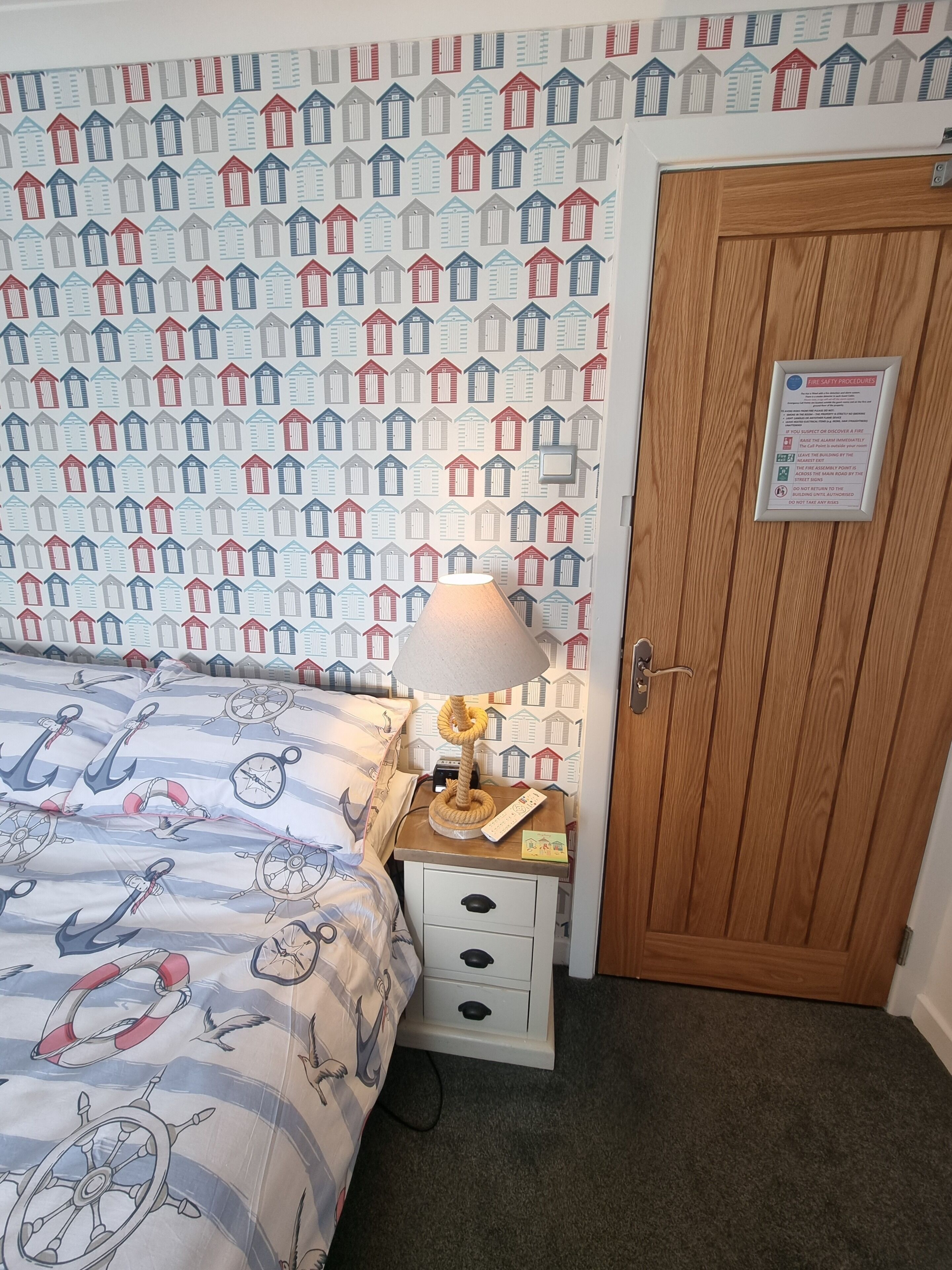 A bedroom with a patterned wallpaper, a bed with nautical-themed bedding, a bedside lamp, a wooden door, and a fire safety sign.