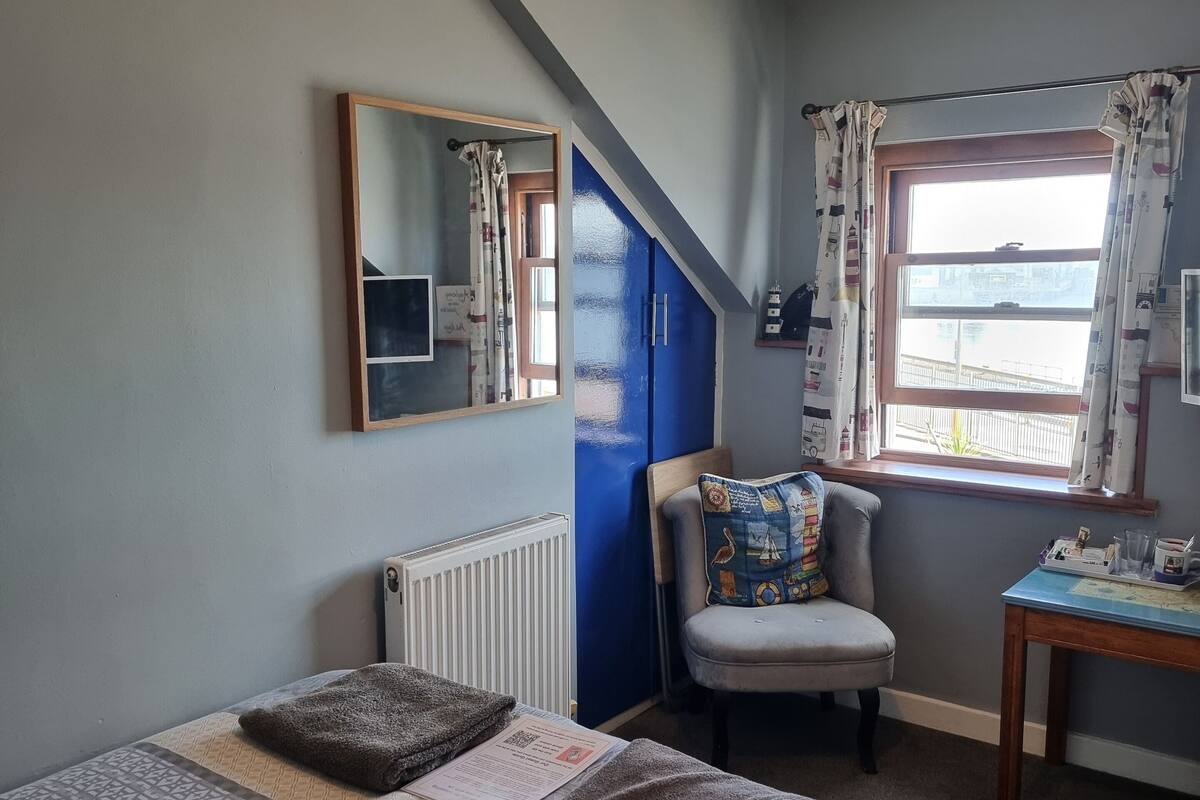 A bedroom with a bed, a chair, a table, a window with curtains, a mirror, and a ceiling light.