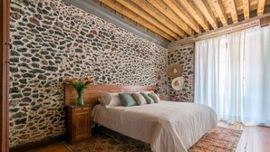Una camera da letto con parete in pietra, travi in legno a soffitto, un letto con testiera, un comodino, un vaso con fiori e un tappeto decorato.