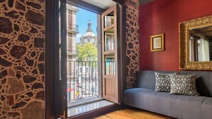 Una habitación con pared de piedra, suelo de madera, sofá gris con cojines estampados, un espejo grande con marco dorado, y vista a un edificio con cúpula a través de una puerta abierta.