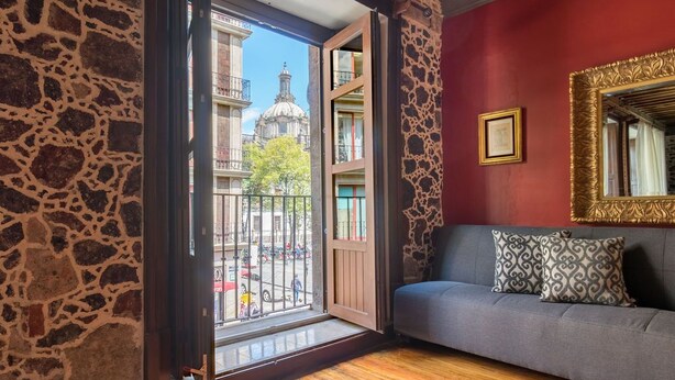 Una stanza con parete in pietra, pavimento in legno, divano grigio con cuscini decorati e un ampio specchio con cornice dorata, oltre a vista su un edificio con una cupola attraverso una porta aperta.