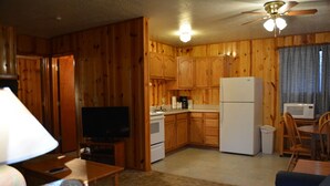 A compact kitchen with wooden cabinets, a refrigerator, a microwave, and a dining area with a table and chairs. - Whispering Pines - Cabin 31 (Spa Tub) (Pinetop)