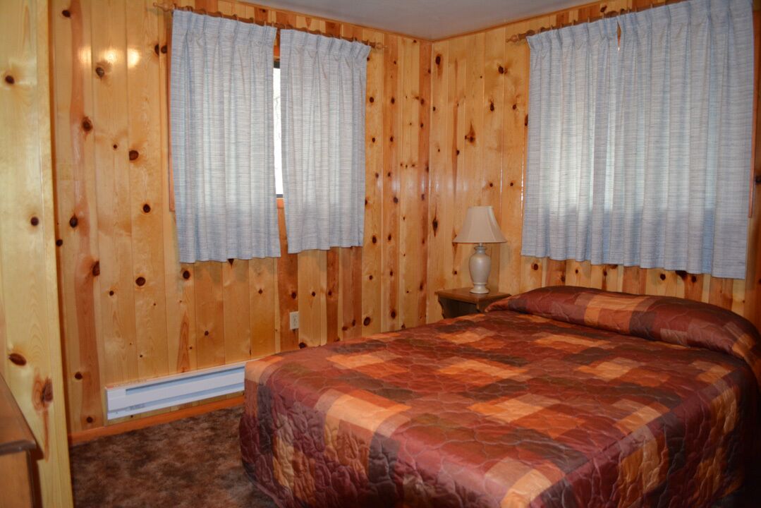 A wooden-paneled room with a bed, two windows with curtains, and a bedside lamp.