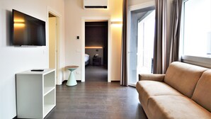 Ein modernes Hotelzimmer mit einer Couch, einem an der Wand montierten Fernseher, einem kleinen Beistelltisch und einem Kleiderschrank mit einem Bett im Hintergrund.