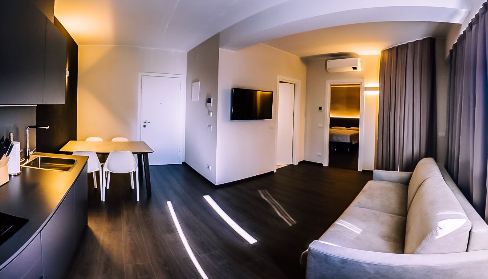 Una sala de estar moderna con un sofá, un comedor con una mesa y sillas, una cocina con un fregadero y armarios, y un televisor montado en la pared.