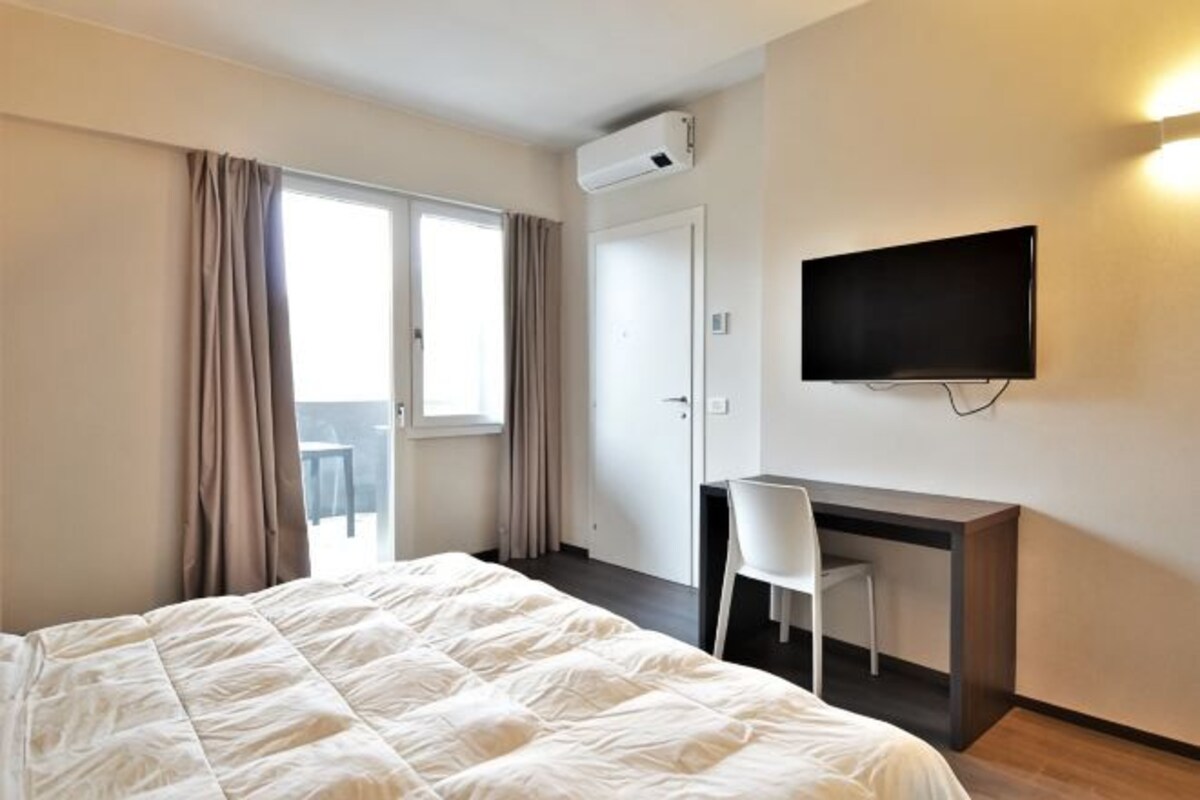 A hotel room with a bed, a desk, a chair, a TV, and an air conditioning unit.