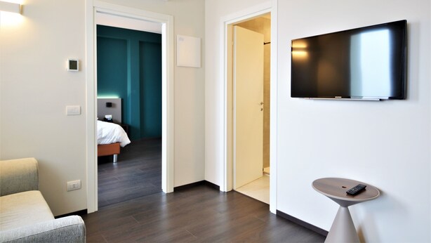 Ein modernes Hotelzimmer mit Flachbildfernseher an der Wand, einem kleinen Tisch mit Fernbedienung und einem Bett mit weißem Kopfteil.