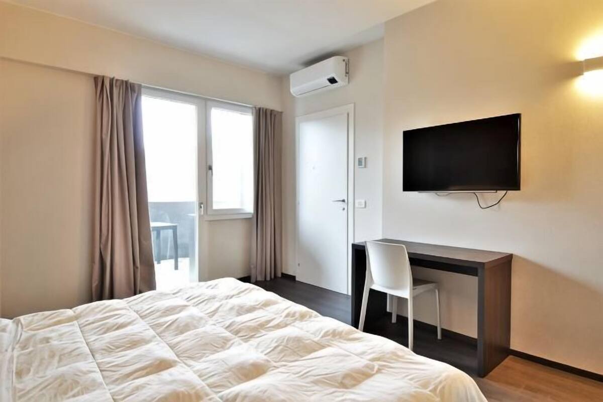 A hotel room with a bed, a desk, a chair, a TV, and an air conditioning unit.