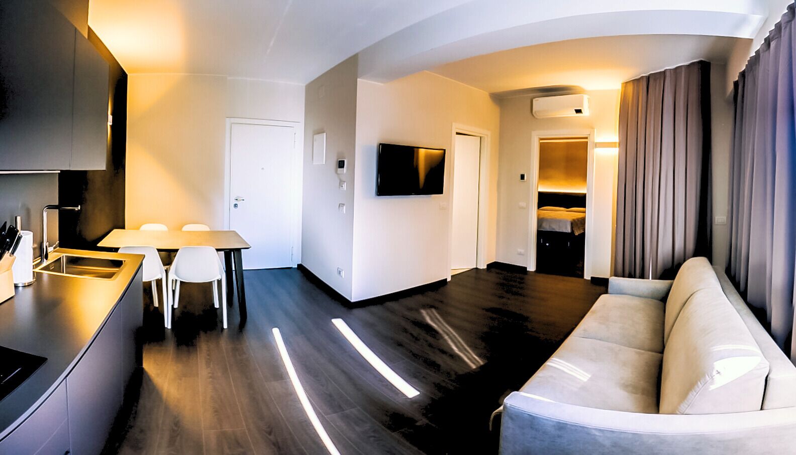 Una sala de estar moderna con un sofá, un televisor montado en la pared y una cocina con una mesa de comedor y sillas.