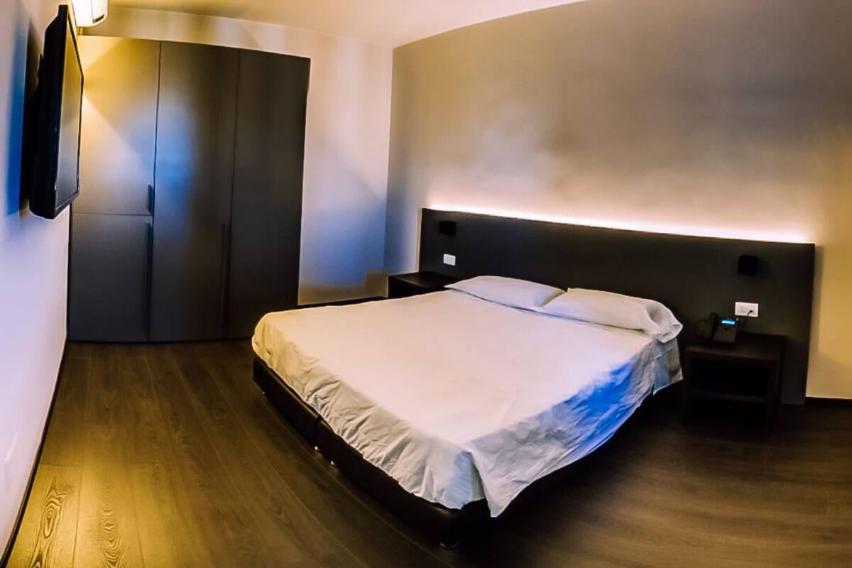 A modern hotel room with a large bed, a bedside table, a built-in wardrobe, and a wall-mounted TV.