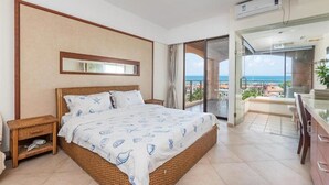 Une chambre avec un grand lit, une table de chevet, un miroir et une vue sur la mer, visible à travers une porte vitrée.