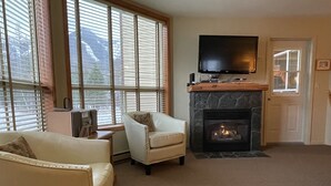 Un salon doté d’une cheminée, d’un téléviseur fixé au-dessus et d’une grande fenêtre avec des stores.