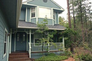 A two-story house with a front porch, a dark blue door, and a small garden area with a pathway leading to the door. - Whispering Pines - Victorian House (Pinetop)