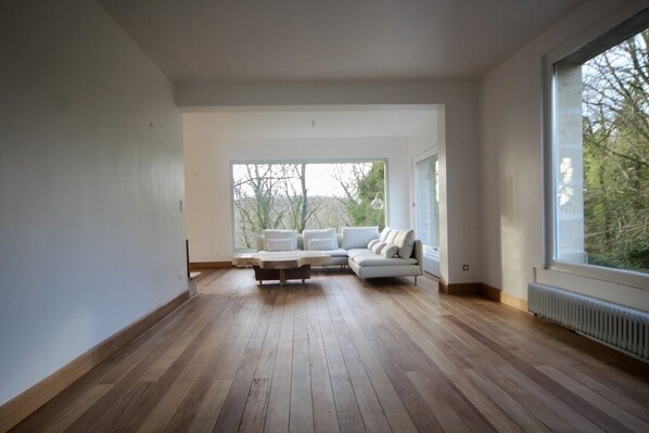 A modern living room with a sectional sofa, wooden flooring, and large windows offering a view of trees.