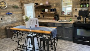 Une cuisine rustique aux murs en bois, avec une table en bois recouverte d’une nappe bleue, une machine à café et un poêle.