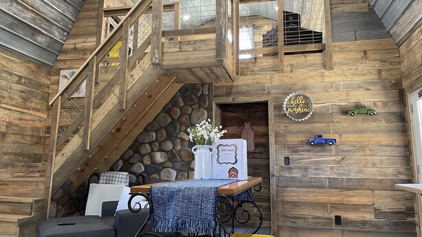 Un intérieur rustique avec un escalier en bois, un mur en pierre, une table recouverte d’une nappe bleue et une pancarte sur laquelle on peut lire « Bonjour, soleil ».