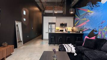 Un intérieur moderne avec un salon aux couleurs sombres, une cuisine dotée d’un îlot central et une fresque murale abstraite et colorée.