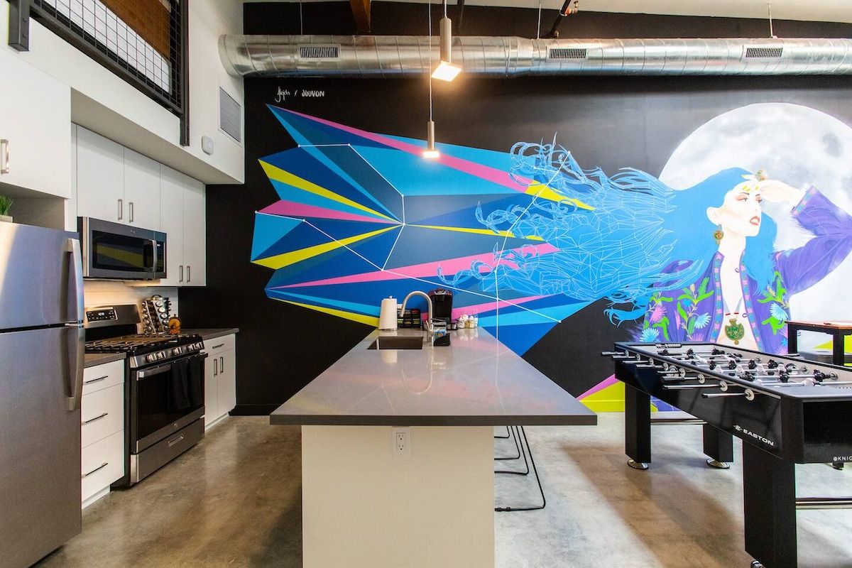 A modern kitchen with a central island, a foosball table, and a large abstract mural on the wall.