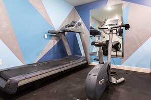 Un gimnasio con una cinta de correr y una máquina elíptica, un espejo que refleja el equipo, y una pared con un diseño geométrico.
