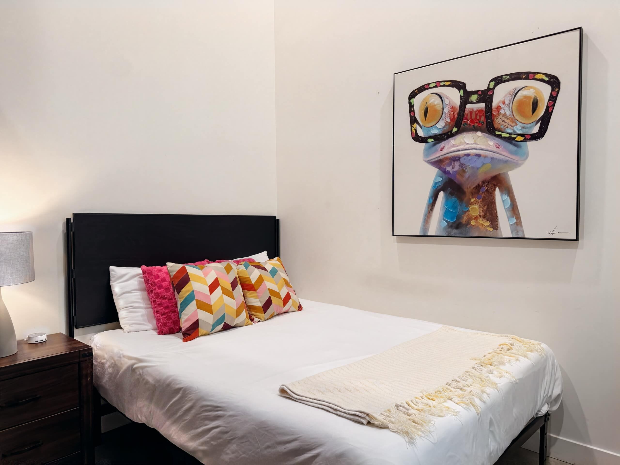 Une chambre à coucher avec un lit, des oreillers, une table de chevet, une lampe et un tableau accroché au mur.