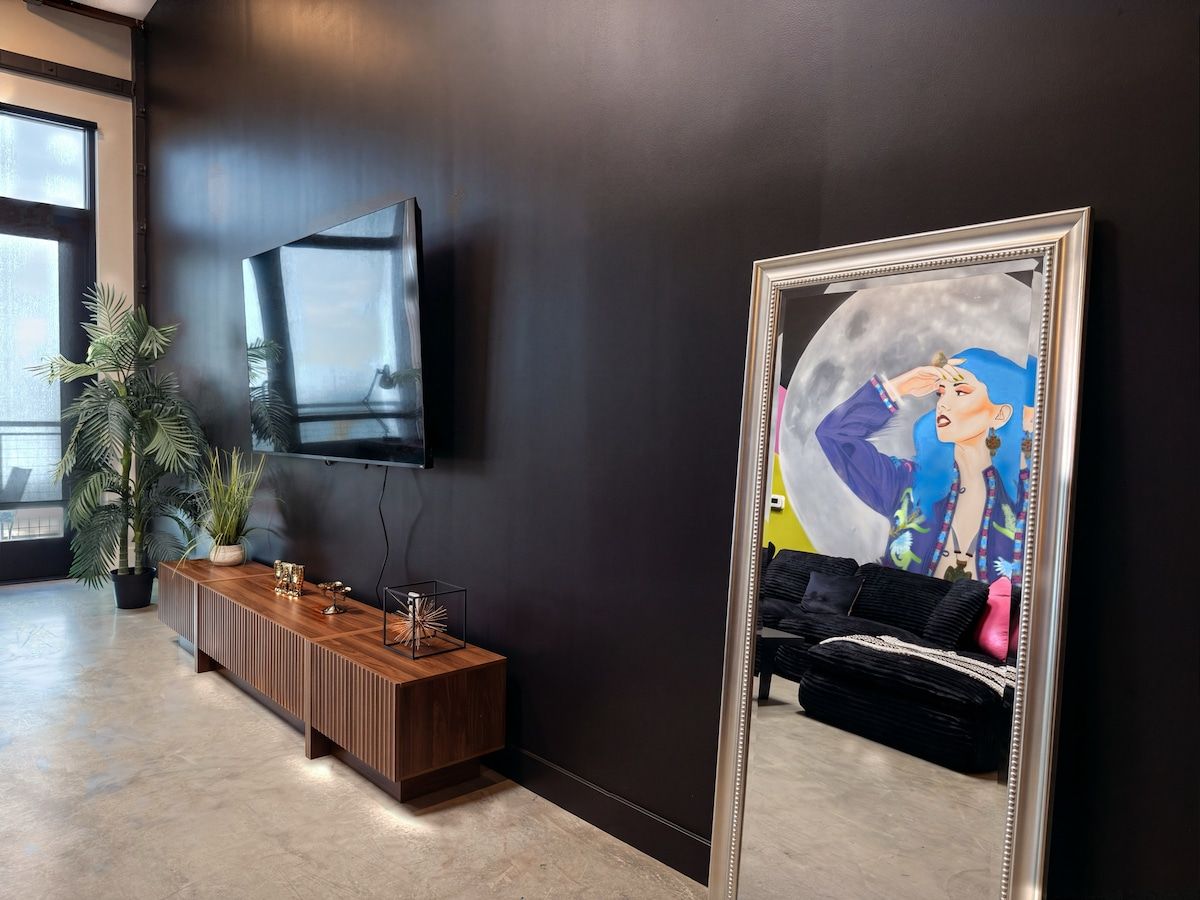 Un interior moderno con una pared oscura, un televisor montado, una consola de madera y un espejo grande que refleja un espacio con un sofá y obras de arte.