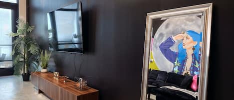 Un interior moderno con una pared oscura, un televisor montado, una consola de madera y un espejo grande que refleja un espacio con un sofá y obras de arte.