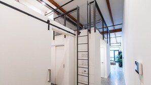 Un pasillo estrecho con una escalera metálica que lleva a un altillo, una puerta con un letrero y una vista a un cuarto con plantas.