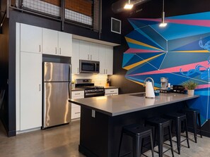 A modern kitchen with stainless steel appliances, a large island with bar stools, and a colorful geometric wall mural. - Stylish Downtown Loft Near Disney (Santa Ana)