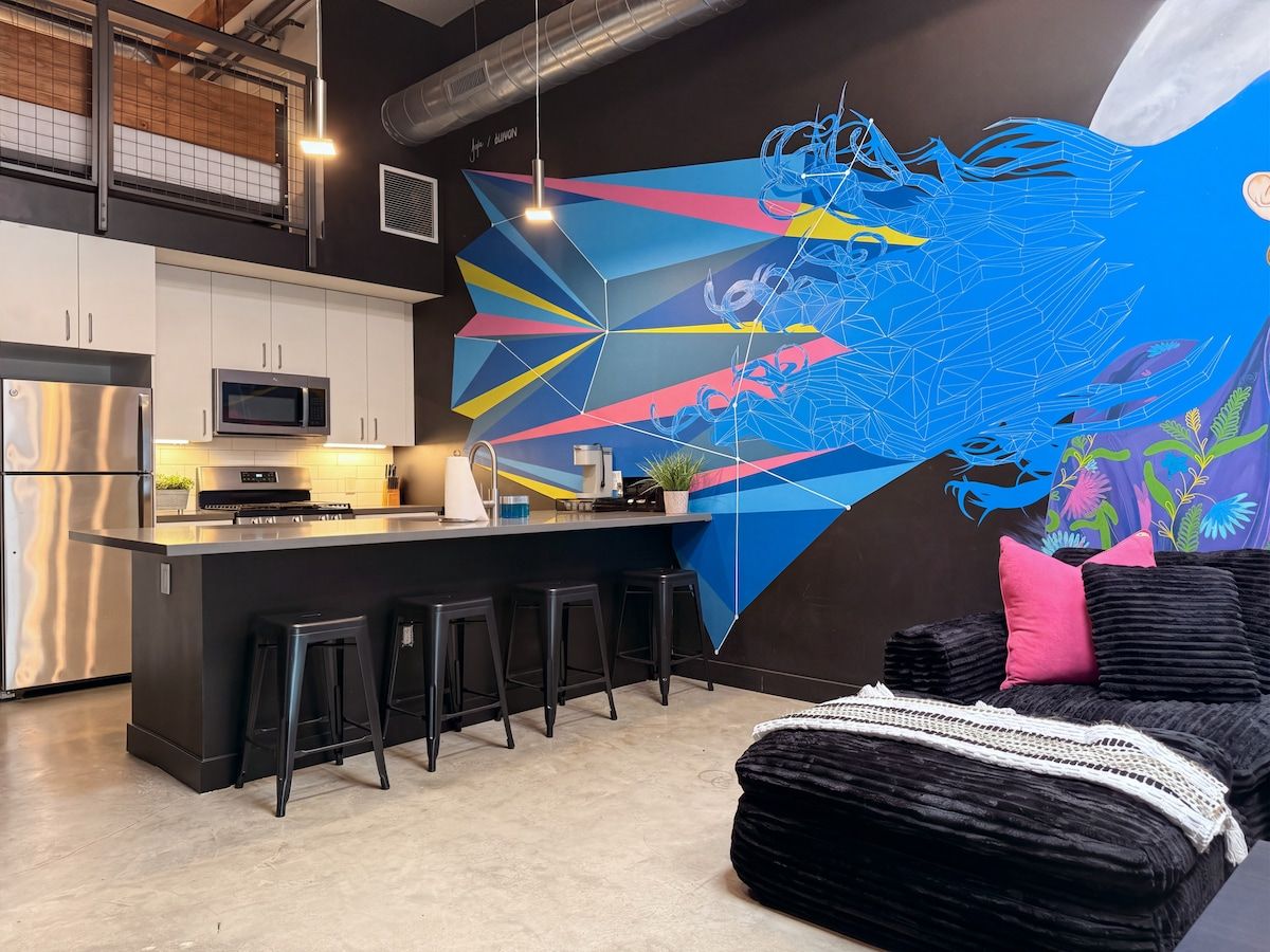 A modern kitchen with a bar area, a large abstract mural, and a black sofa with a white throw.