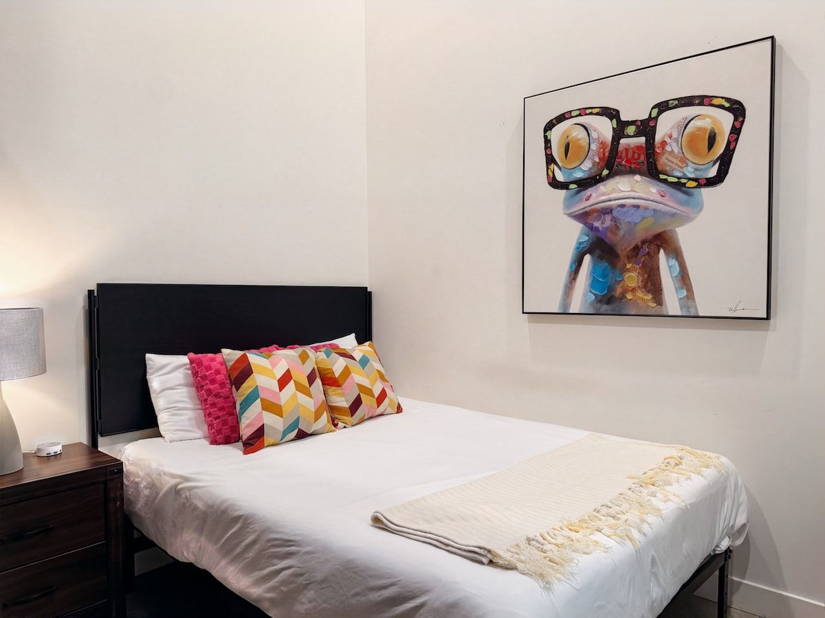 A bedroom with a bed, pillows, a nightstand, a lamp, and a wall-mounted artwork.