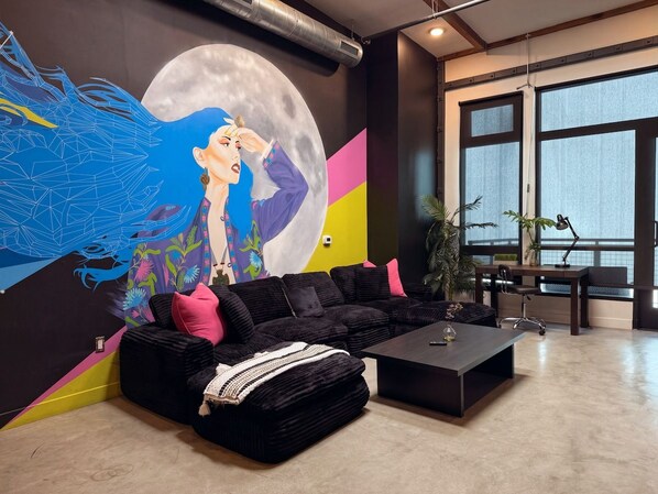 A modern living room with a large wall mural, a black sectional sofa, a coffee table, and a desk with a chair.