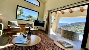 Una sala de estar moderna con un televisor grande, asientos cómodos, una mesa de centro y vistas a un paisaje montañoso a través de amplios ventanales.