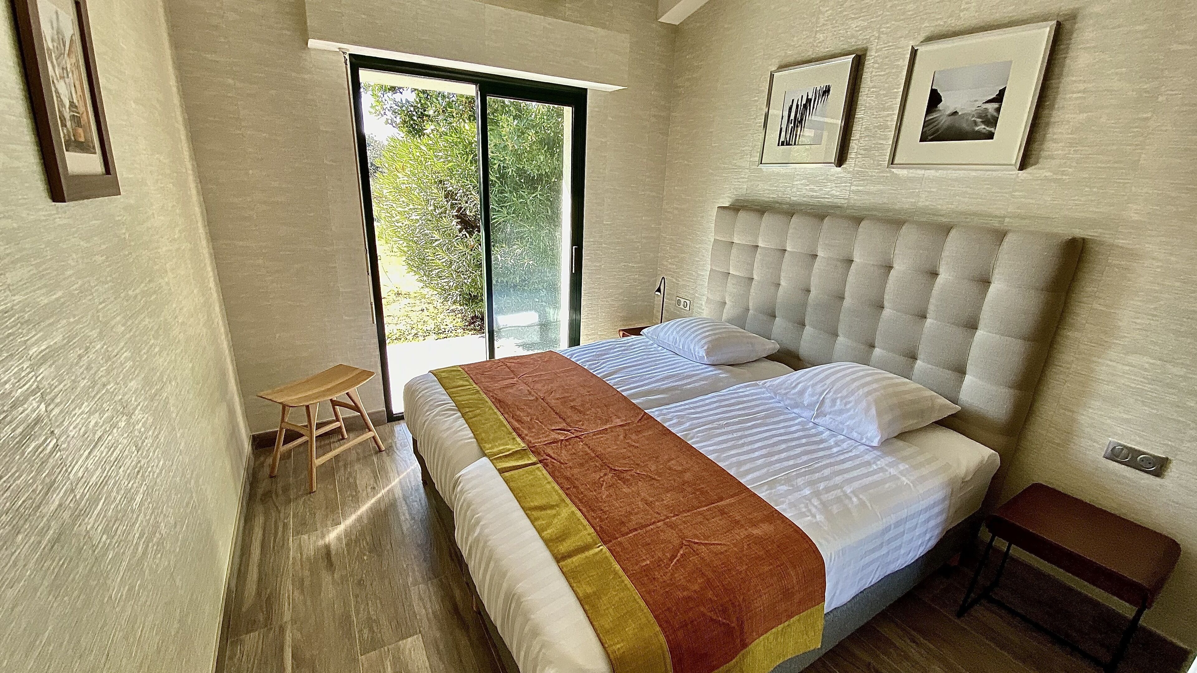 Un dormitorio con una cama grande, un taburete pequeño, una mesita de noche y un ventanal corredero que da a un balcón.