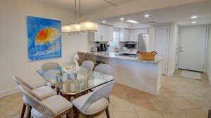 Une cuisine moderne avec des meubles blancs, un îlot central et un coin repas comprenant une table en verre et des chaises en osier.