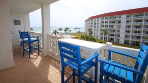 Un balcon avec des chaises bleues et une table blanche, donnant sur une plage et un bâtiment d’hôtel.