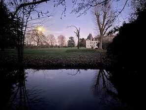 A large, historic building with a symmetrical facade and a prominent tower, surrounded by a well-maintained lawn and a calm water body reflecting the structure.