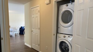 Un lavadero con dos lavadoras de carga frontal apiladas, una zona de comedor con una silla azul y una puerta blanca.
