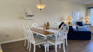 Un comedor con mesa de madera y sillas blancas, una lámpara colgante y un estante con un modelo de velero y un pájaro.