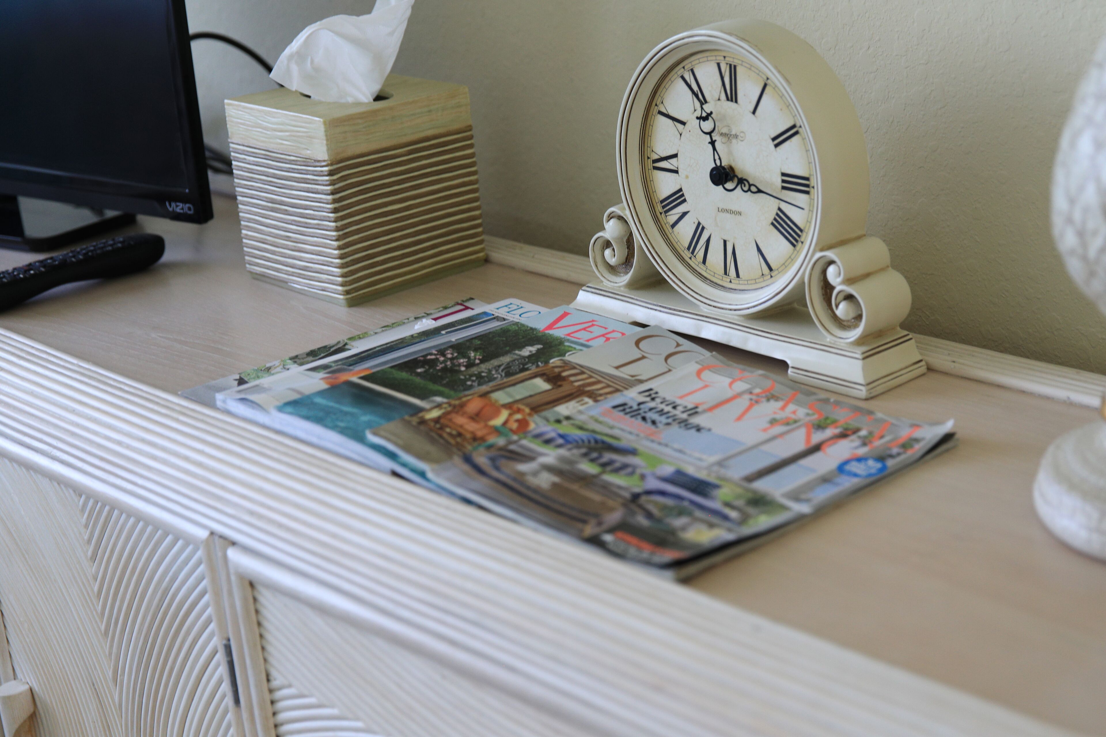 A desk with a clock, a remote control, and a stack of magazines.