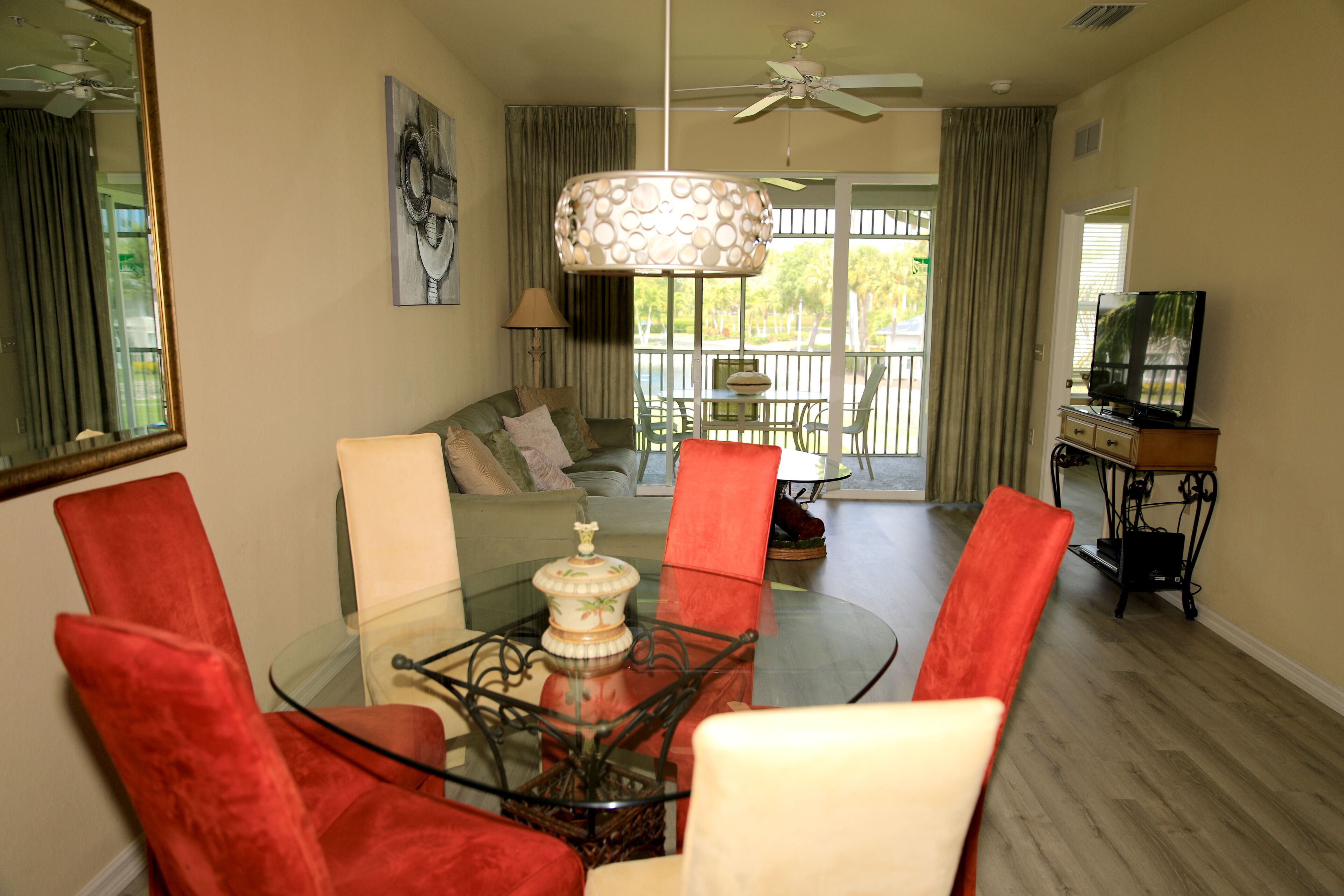 A dining area with a glass table, red chairs, a ceiling fan, and a television in the background.