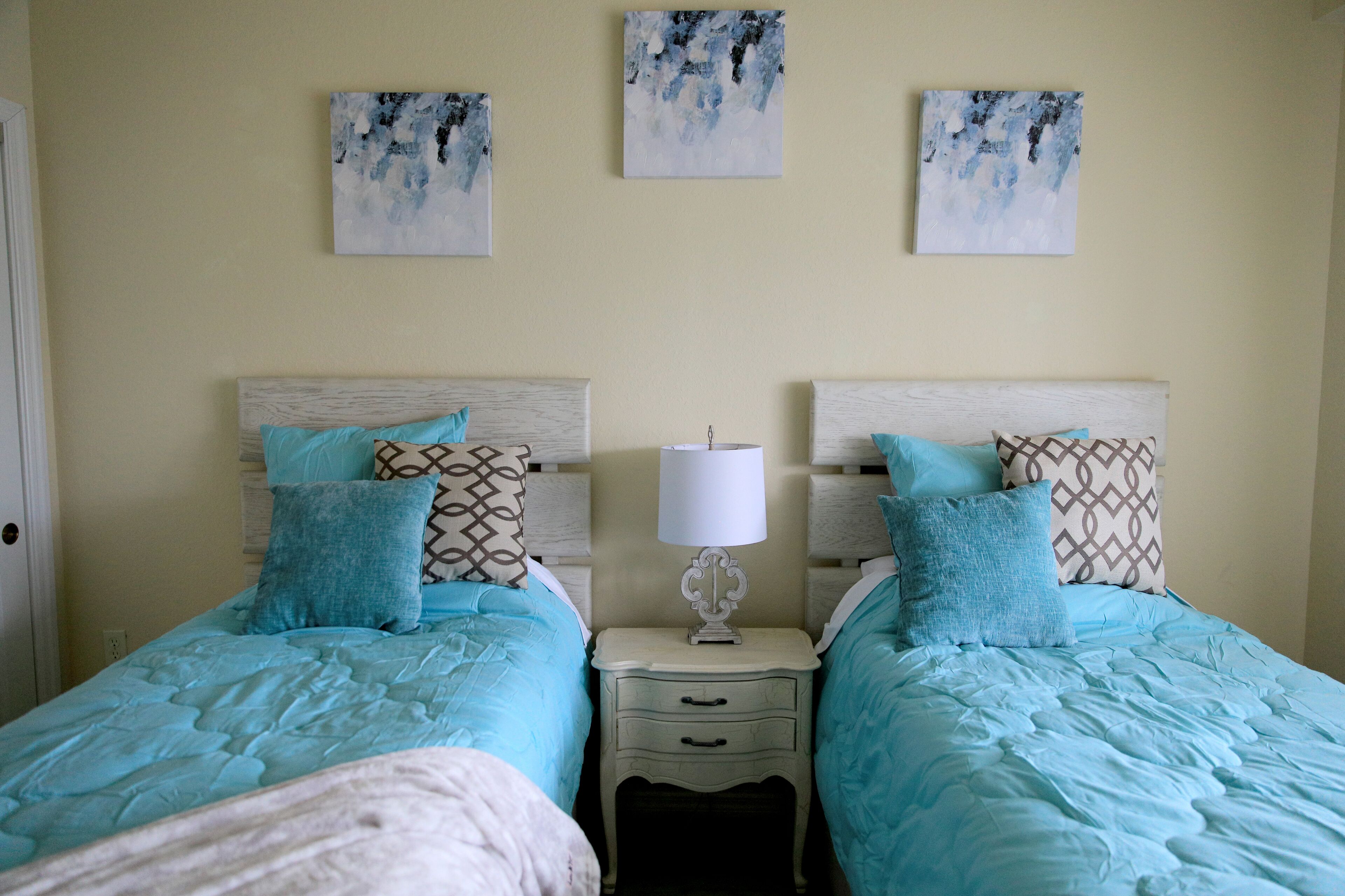 Une chambre à coucher avec deux lits, chacun doté d’une tête de lit, d’une table de chevet avec une lampe, et de trois tableaux accrochés au mur.