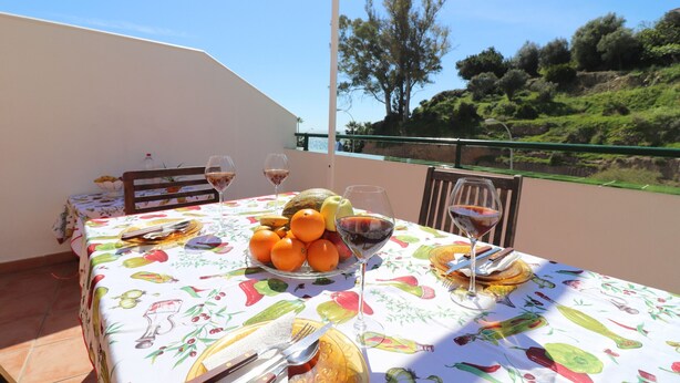 Mesa posta com frutas, copos de vinho e talheres numa varanda com vista para árvores e colinas.