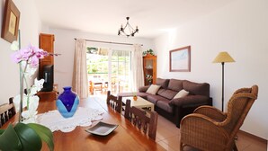 Uma sala de estar com sofá, mesa de jantar, cadeiras, um vaso de flores, televisão e uma janela com cortinas.